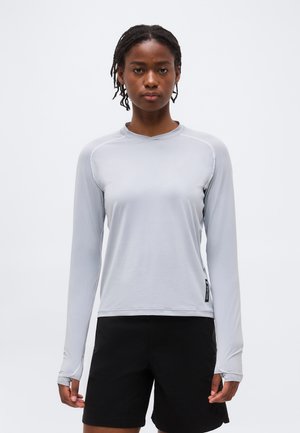 Femme aux cheveux tressés portant un haut de sport gris clair à manches longues et un short noir, posant devant un fond blanc uni.