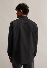 Črna dolga majica s dolgimi rokavi, ravnega kroja, z gumbnimi manšetami in zanko na hrbtni strani. Gladka tekstura in minimalističen dizajn.