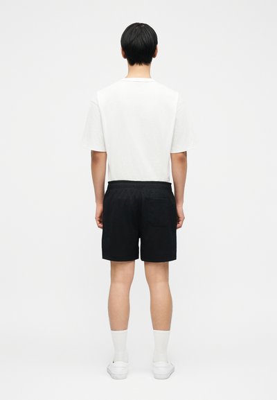 Άνδρας που στέκεται με την πλάτη, φοράει λευκό μπλουζάκι, μαύρο σορτς με πίσω τσέπη, λευκές κάλτσες και λευκά αθλητικά παπούτσια σε απλό φόντο.