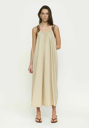 Naine kannab lahtist, beežikat varrukateta maksikleit pimendatud õlapaeltega ja pruune sandaale, seistes ühetaolise valge tausta ees.