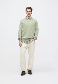 Pulôver de malha verde claro sobre uma camisa cinza riscada, combinado com calças largas de tom creme e sapatos escuros de slip-on. Linhas simples e limpas.