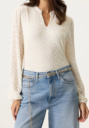 Valkoinen pitsipaita pitkällä hihalla, jossa siksak-kuvio, ja siniset korkeavyötäröiset farkut, asusteena kultainen ketjuvyö, joka roikkuu löysästi.