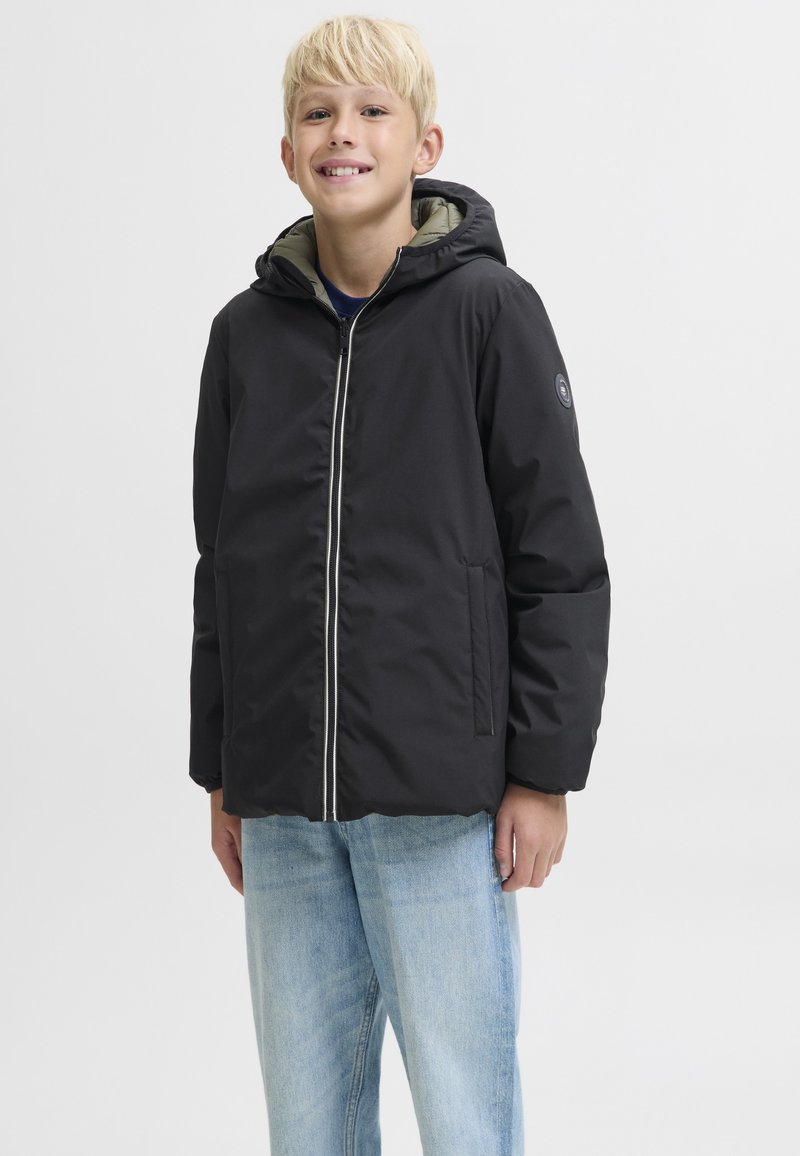 Niño rubio sonriente con chaqueta negra con capucha y cremallera y vaqueros azul claro, de pie contra un fondo sencillo.