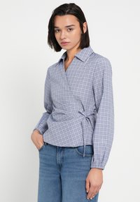 Μπλούζα τύπου wrap σε ανοιχτό μπλε ύφασμα με καρό σχέδιο. Διαθέτει γιακά, μακριά μανίκια και λεπτομέρεια δεσίματος στη μέση. Συνδυασμένη με τζιν.