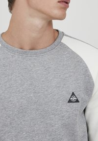 Grå sweatshirt med raglan-hvide ærmer, ribbet halsudskæring og et sort trekantet logo-patch med ordet "BLEND." Blødt, tekstureret stof.