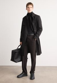 Sort trenchcoat over en sort sweater med høj hals, parret med mørkebrune bukser og sorte sneakers. Holder en struktureret sort taske.