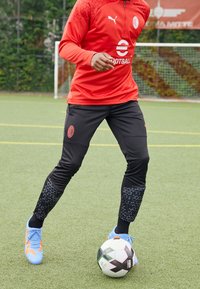 Czerwony długi rękaw sportowy top z teksturowanymi ramionami, czarne spodnie z wzorzystymi dolnymi nogawkami, niebieskie korki do piłki nożnej i czarno-biała piłka nożna.