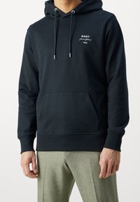 Felpa nera con cappuccio, con cordini, tasca a marsupio e logo GANT in bianco. Realizzata in morbido cotone, con polsini e orlo a costine per un design sagomato.