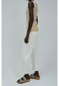 Donkerhuidige persoon die een beige gebreide mouwloze top, witte cropped wijde jeans en beige suede sandalen met twee gespen draagt.