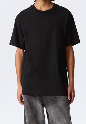 Mężczyzna ubrany w gładki czarny t-shirt z krótkim rękawem i wyblakłe szare dżinsy, stojący z rękami swobodnie opuszczonymi wzdłuż ciała, na białym tle.