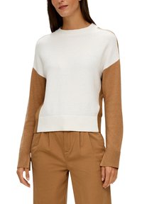 Femme portant un pull à blocs de couleurs avec un haut blanc, des manches marron et un pantalon marron assorti, se tenant debout avec les mains détendues le long du corps.