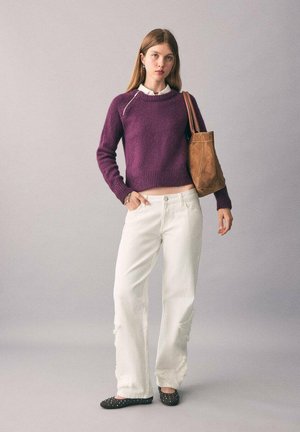 Pull en tricot violet avec ourlet côtelé, jean blanc usé et sac fourre-tout en cuir marron. Des chaussures plates sombres à clous complètent la tenue.