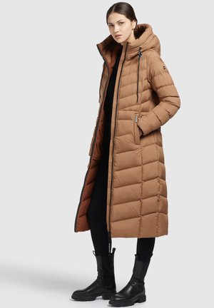 Manteau long et marron en duvet avec capuche, présentant une texture matelassée horizontale, une fermeture éclair à l'avant et des poches latérales, porté sur une tenue noire.