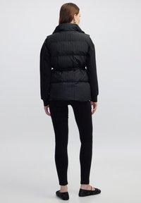 Frau in einer schwarzen gesteppte Weste über einem schwarzen Langarmshirt, schwarzen Leggings und schwarzen verzierten Ballerinas, steht mit dem Rücken zur Kamera.