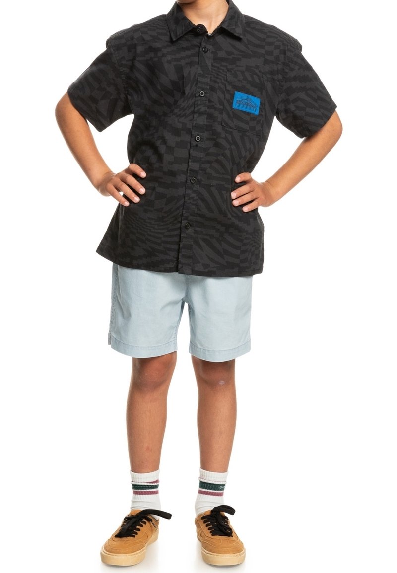 Enfant debout, les mains sur les hanches, portant une chemise noire à manches courtes à motifs avec boutons, un short bleu clair, des chaussettes blanches et des baskets beige avec lacets noirs.