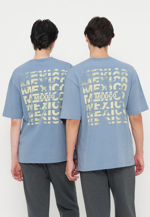 Due persone che indossano magliette blu con testo giallo sbiadito che ripete "Mexico" sulla schiena e pantaloni grigio scuro, in piedi fianco a fianco.