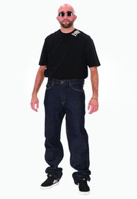 T-shirt noir avec logo, jean bleu foncé ample et baskets noires avec des accents blancs. Des lunettes de soleil rondes complètent le look.