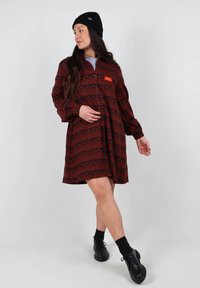 Robe à manches longues, de couleur rouge avec des motifs géométriques, à boutons sur le devant et col rond ; associée à un bonnet en tricot noir et des chaussures.