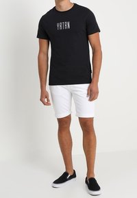 Muž oblečený v černém tričku YRTN, bílých krátkých kalhotách a černých botách na slip-on, stojí proti jednoduchému světlému pozadí.