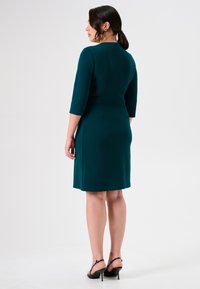 Türkises knieumspielendes Kleid mit dreiviertel Ärmeln und taillierter Taille, von hinten präsentiert. Model trägt schwarze High-Heels.
