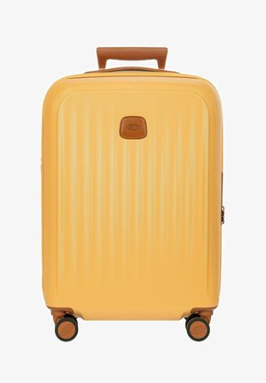 Valise rigide jaune avec poignée marron et quatre roues, avec un petit patch logo en cuir marron au centre.