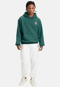 Felpa verde con cappuccio, tasca frontale e patch con logo, abbinata a pantaloni della tuta bianchi e sneakers color crema. Tessuto strutturato, vestibilità comoda.