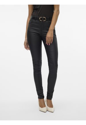 Vrouw die zwarte nauwsluitende leren broek draagt met een zwarte riem en witte hoge hakken, staand tegen een witte achtergrond.