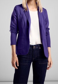 Fioletowa marynarka z elastycznej tkaniny z długimi rękawami, z dwoma przednimi kieszeniami, noszona na białej bluzce i ciemnoniebieskich dżinsach.