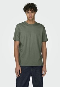 Camiseta verde oliva de manga corta hecha de algodón, con cuello redondo y corte relajado. Jeans con acabado de mezclilla oscura visibles por debajo.