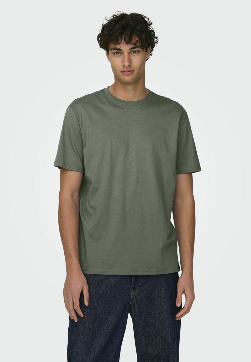 Camiseta verde oliva de manga corta hecha de algodón, con cuello redondo y corte relajado. Jeans con acabado de mezclilla oscura visibles por debajo.