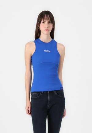 Blå ribbet tanktopp med høy hals og tettsittende design. Har hvit tekst "RACING" på brystet, sammen med mørke jeans.