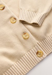 Cardigan beige en tricot avec poignets et ourlet côtelés, doté de six boutons ronds crème à finition brillante.