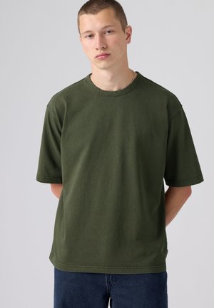 Giovane uomo con capelli corti che indossa una maglietta oversize verde oliva semplice e pantaloni blu scuro davanti a uno sfondo chiaro e uniforme.