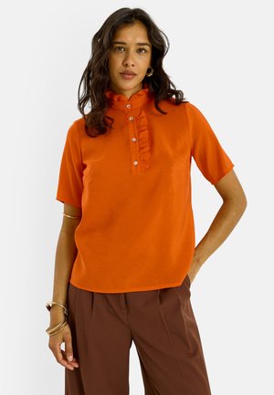 Mujer con cabello oscuro que lleva una blusa naranja de manga corta con tapeta de botones fruncida y pantalones marrones plisados, posando con la mano en la cadera.