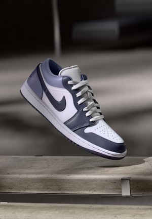 Weißer und grauer Nike Air Jordan Sneaker mit schwarzem Swoosh, weißen Schnürsenkeln, schwebend über einer Betonoberfläche bei weichem Licht.