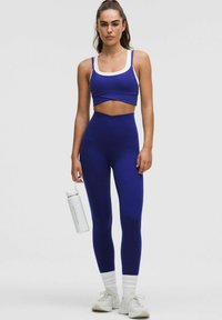Soutien-gorge de sport violet avec des bordures blanches et des leggings taille haute assortis. La modèle tient une bouteille d'eau transparente, portant des baskets et des chaussettes blanches.
