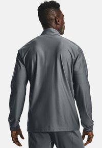 Giacca sportiva grigia con colletto, maniche lunghe e polsini elasticizzati. Tessuto liscio e design affusolato. Senza loghi o motivi visibili.