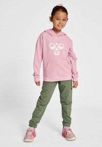 Rosa Kapuzenpullover mit weißem Grafikdesign, Fronttasche, kombiniert mit grünen Jogginghosen und rosa Sportschuhen mit weißen Akzenten.
