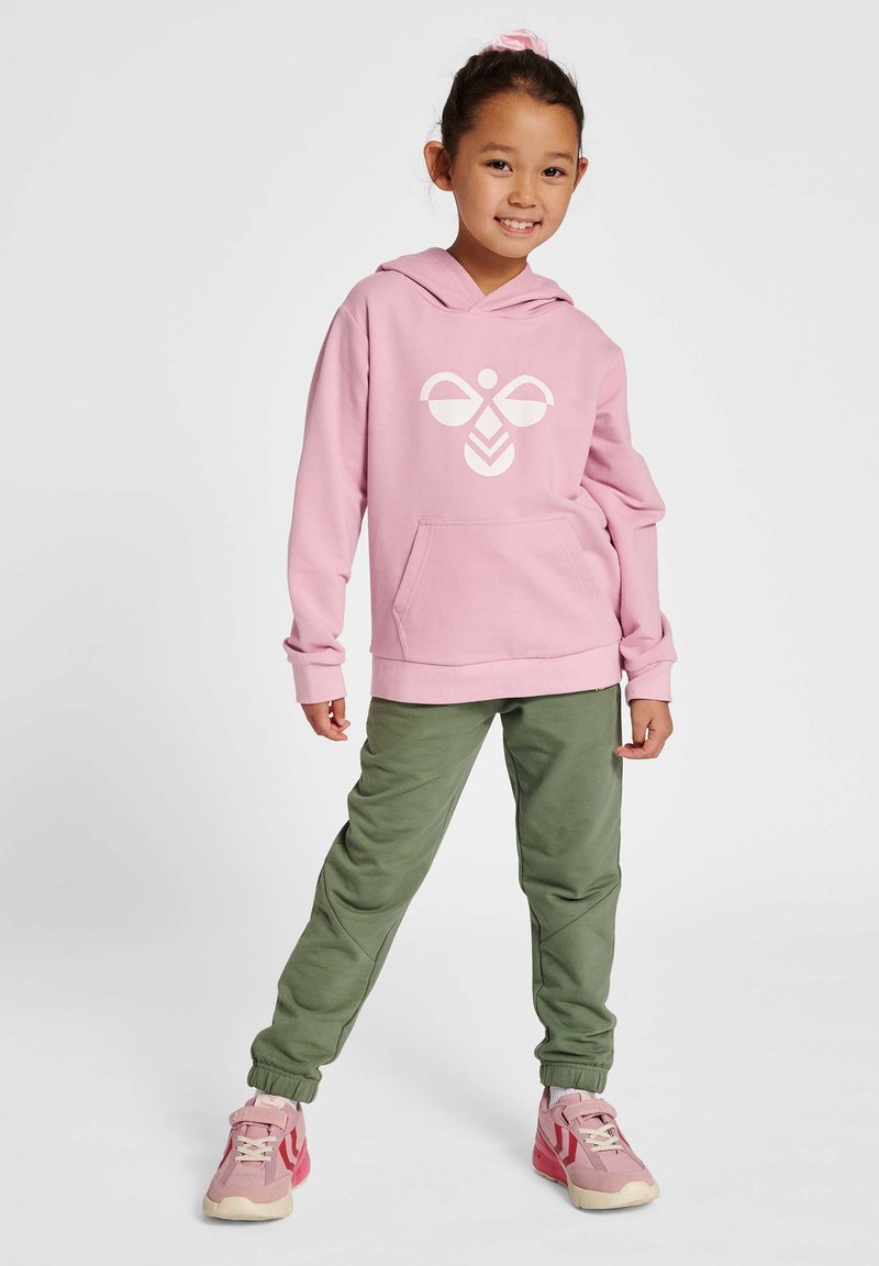 Rosa Kapuzenpullover mit weißem Grafikdesign, Fronttasche, kombiniert mit grünen Jogginghosen und rosa Sportschuhen mit weißen Akzenten.