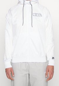 Bílá mikina na zip s kapucí, černými šňůrkami a šedým logem Nike na hrudi; elastické manžety a pas, hladká textura.