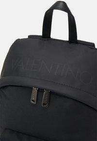 Mochila negra de nylon con una textura suave, un asa superior y cremalleras mate con la marca "Valentino" en la parte frontal.