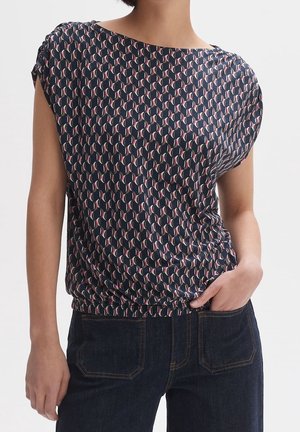 Chemisier marine avec un motif circulaire en rose et crème, manches courtes et ourlet froncé. Associé à un jean en denim foncé.