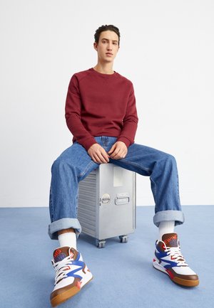 Roter Pullover, blaue Jeans mit umgekrempelten Säumen und weiße/blau/braune Sneaker. Sitzend auf einer silbernen Aufbewahrungsbox, schlichter Hintergrund.