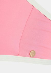 Rozā bikini tops ar balto apdari, gludu tekstūru un nelielu zelta metāla logo akcentu tuvumā šuvēm.