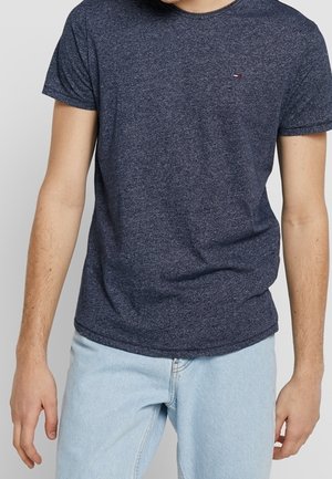 Mężczyzna w ciemnoniebieskim t-shircie z krótkim rękawem i jasnoniebieskich dżinsach, stojący z rozluźnionymi ramionami wzdłuż ciała.