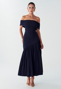 Femme portant une robe midi bleue marine épaules dénudées avec détail volant asymétrique, boucles d'oreilles créoles dorées, bracelet et sandales beiges à talons.