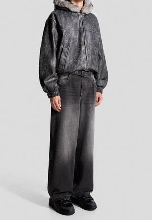 Personne portant une veste zippée gris foncé avec une capuche bordée de fourrure, un jean noir délavé à jambes larges et des baskets noires, debout devant un fond uni.
