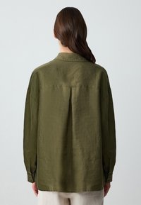 Camicia a maniche lunghe verde oliva realizzata in tessuto testurizzato. Presenta un colletto, polsini con bottone e una piega sul retro, con una vestibilità ampia.