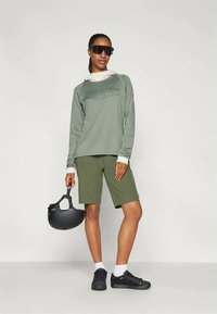 Γυναίκα με πράσινο πουλόβερ μακρύ μανίκι και σορτς, μαύρα γυαλιά ηλίου, λευκές κάλτσες, μαύρα αθλητικά παπούτσια, κρατώντας ένα μαύρο κράνος ποδηλάτου στο πλάι της.