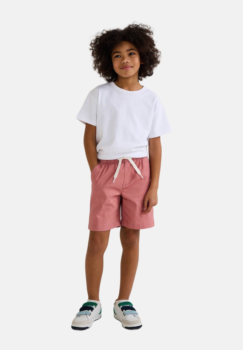 T-shirt blanc à manches courtes, short rose avec cordon de serrage, et baskets blanches avec accents verts. Design simple, coupe décontractée et tissu léger.